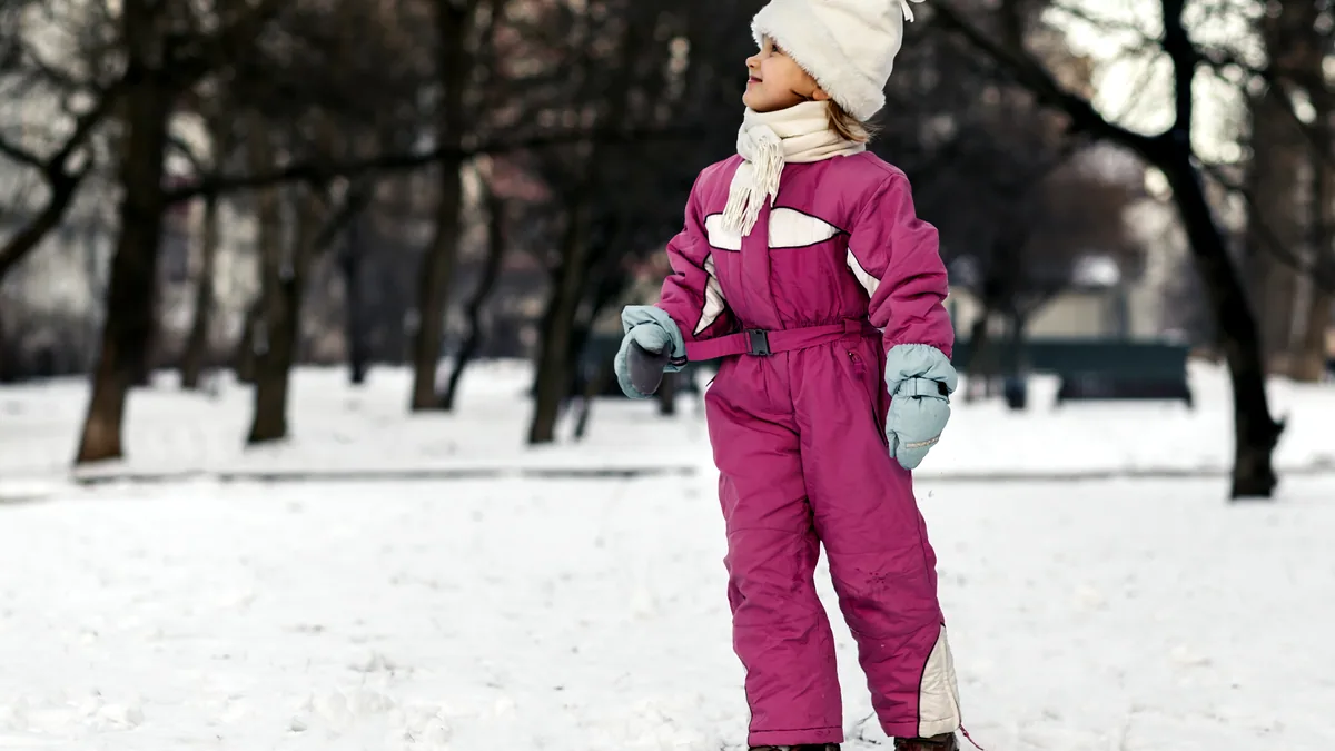 A child in a puffy snowsuit standing in deep snow, breath visible in the cold air.