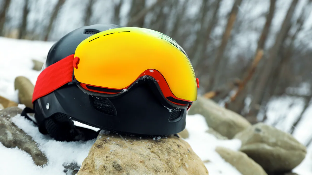 A matte black snowboard helmet resting on a rocky outcrop with a blurred alpine backdrop.