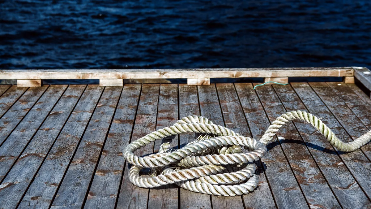 A coiled waterski rope and float lying on a weathered wooden dock beside still water.